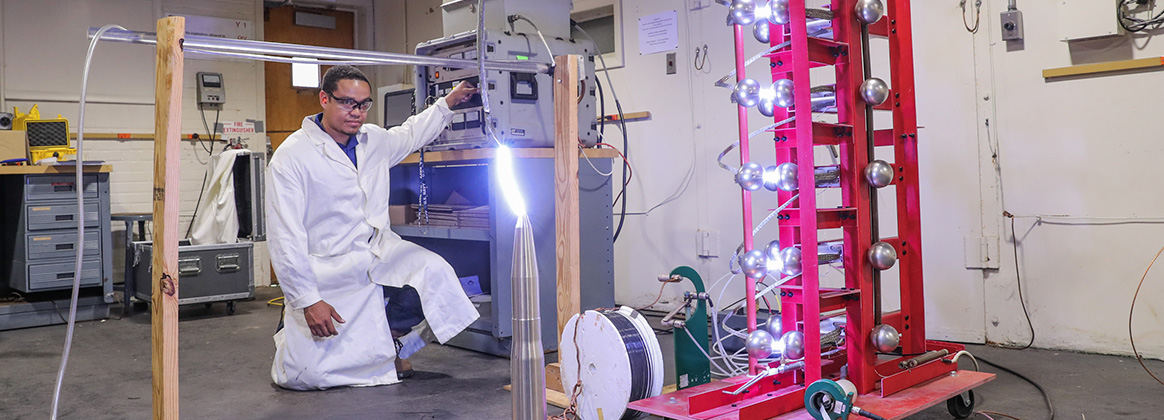 Naval Surface Warfare Center Indian Head Division Systems Engineering Department ElectroStatic Discharge (ESD) Tech Agent Team Lead Phillip Melton observes the transient arc of 300kV to the discharge point between the Helicopter-ESD (HESD) Marx and the INERT ordnance on the ground plane during Bare Ordnance HESD testing. 