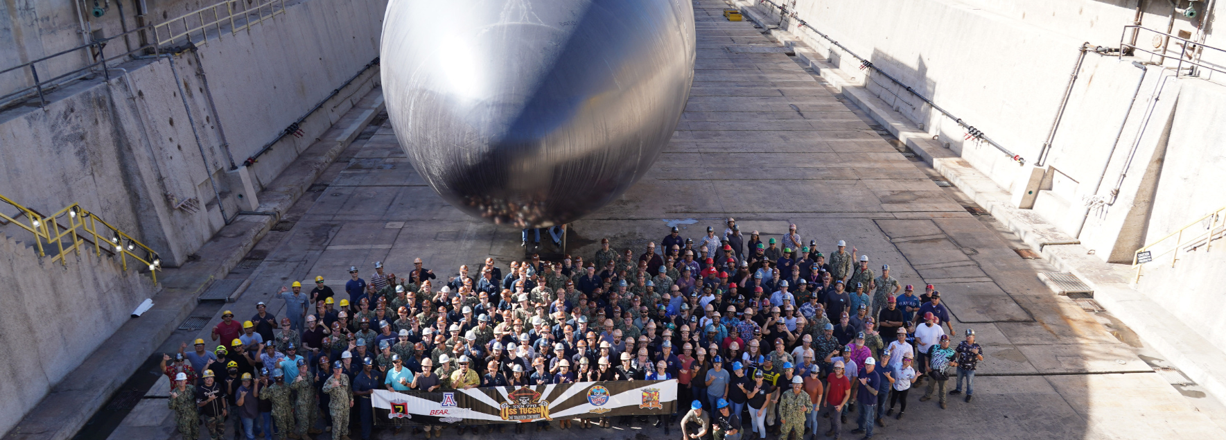 People stand with a banner in front of submarine USS Tucson in a dry dock at Perl Harbor Naval Shipyard